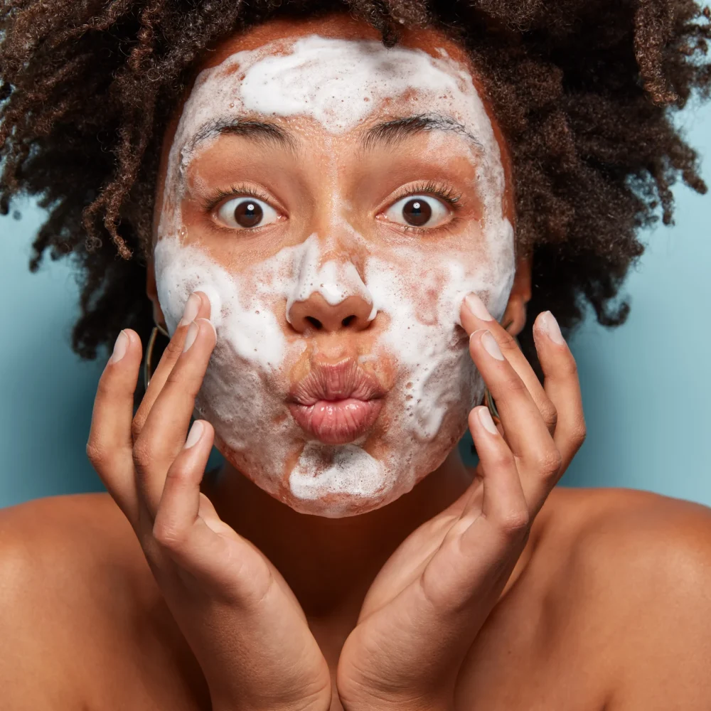 people-wellness-hygiene-skin-problems-concept-lovely-afro-american-lady-keeps-lips-folded-touches-cheeks-has-white-foam-faces-washes-with-beauty-gel-feels-refreshed-has-wide-opened-eyes